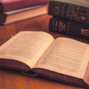 Op de afbeelding: Een open boek met verouderde, vergeelde pagina's en een bruine leren kaft. Het boek ligt open op een houten tafel met andere boeken die erachter gestapeld zijn.