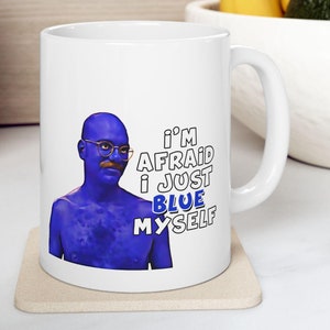 Peut inclure: Tasse en céramique blanche avec une figurine à la peau bleue portant des lunettes et une moustache. La tasse porte l'inscription "I'M AFRAID I JUST BLUE MYSELF" en noir et bleu. La tasse est posée sur un dessous de verre beige.