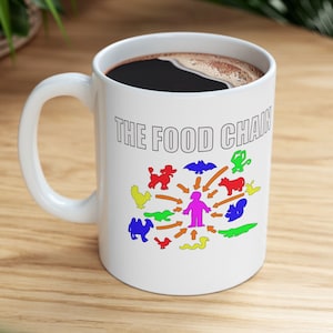 May include: White ceramic mug with a black handle. The mug has a graphic of a food chain with a person at the center and various animals around them. The text "THE FOOD CHAIN" is printed in black at the top of the mug.