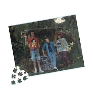 Può includere: Tre persone stanno in piedi davanti a un muro di pietra. La persona a sinistra indossa una camicia rossa con un'immagine di Topolino e jeans blu. La persona al centro indossa una camicia blu e jeans blu. La persona a destra indossa una camicia bianca con il testo "Class of '84" e jeans blu.