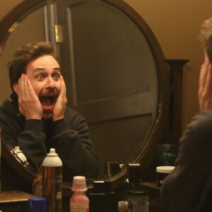 May include: A man wearing a black hooded top looks in the mirror with a surprised expression. He is holding his hands to his face and his mouth is open in shock. There are various toiletries on the counter in front of him.