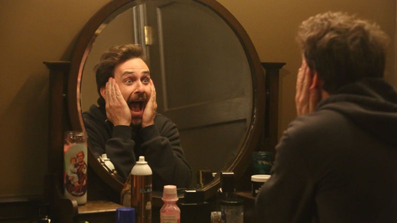 May include: A man wearing a black hooded top looks in the mirror with a surprised expression. He is holding his hands to his face and his mouth is open in shock. There are various toiletries on the counter in front of him.