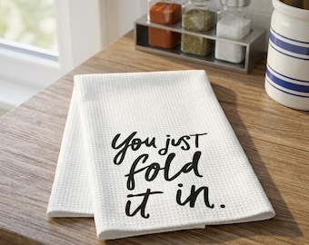 May include: A white waffle-weave kitchen towel featuring the black printed text "You just fold it in." The towel is folded and rests on a wooden surface, with glass spice jars and a blue and white striped ceramic container visible nearby.