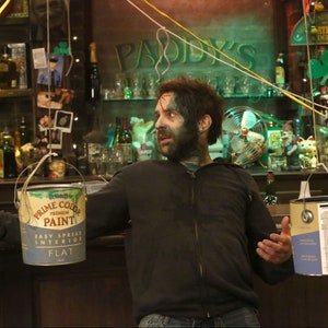 May include: A man with green paint on his face and beard stands in a bar with a paint can hanging from the ceiling. The can is labelled "Prime Color Premium Paint Easy Spread Interior Flat".
