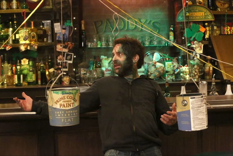 May include: A man with green paint on his face and beard stands in a bar with a paint can hanging from the ceiling. The can is labelled "Prime Color Premium Paint Easy Spread Interior Flat".