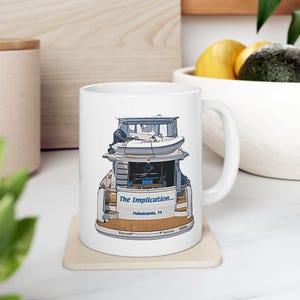May include: White ceramic mug featuring a colourful illustration of a boat with the text "The Implication... Philadelphia, PA" printed on the side.