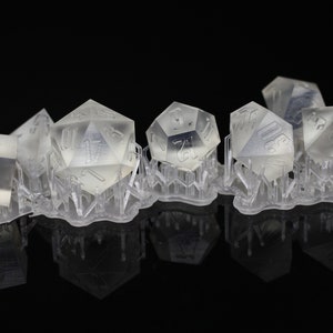 May include: A set of six clear plastic dice with black numbers. The dice are arranged in a row on a black surface. The dice are all different shapes and sizes.