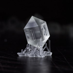 Puede incluir: Un dado de plástico transparente de doce caras con el número 12 impreso en él. El dado está apoyado en una base de plástico transparente con un diseño geométrico.