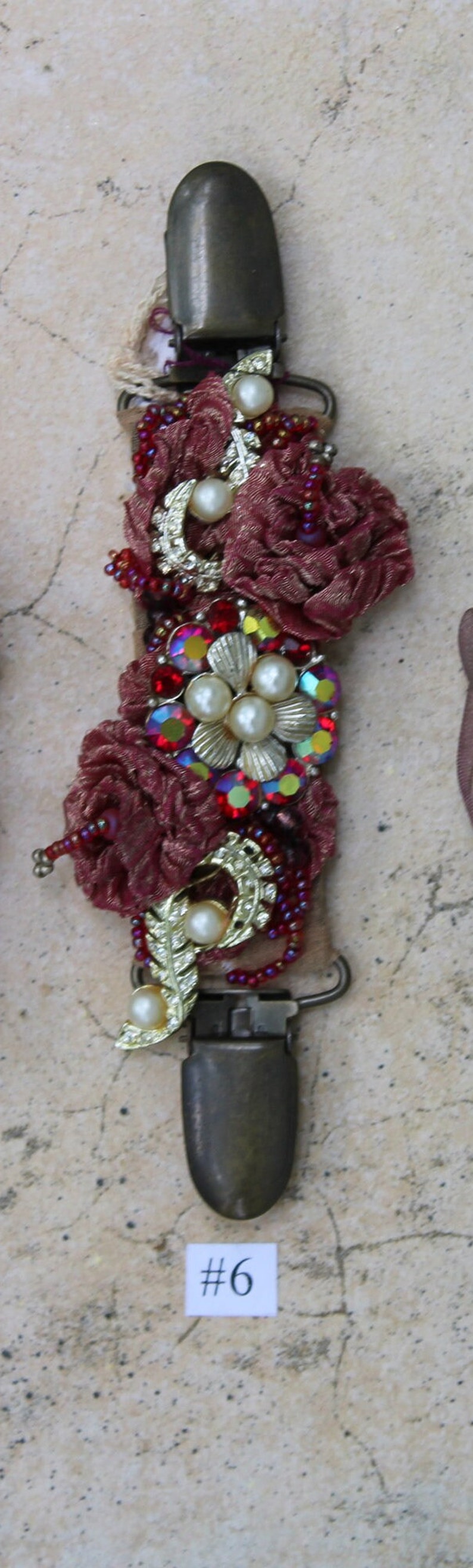 May include: A burgundy fabric flower embellishment with gold and pearl accents, red and iridescent rhinestones, and a metal clasp. The embellishment is labeled with the number "#6".
