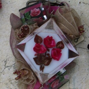 May include: A vintage-style metal clasp with a decorative floral design. The clasp features a white background with pink roses and brown leaves. The clasp is surrounded by brown and pink fabric and beads.