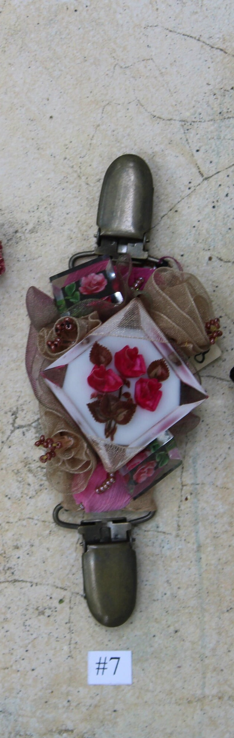 May include: A vintage-style metal clasp with a decorative floral design. The clasp features a white background with pink roses and brown leaves. The clasp is surrounded by brown and pink fabric and beads.