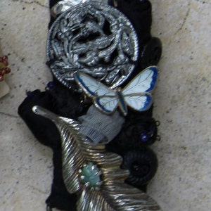 May include: A silver and black metal brooch with a butterfly, leaf, and floral design. The brooch is attached to a black fabric base. The number #8 is printed on a white label below the brooch.