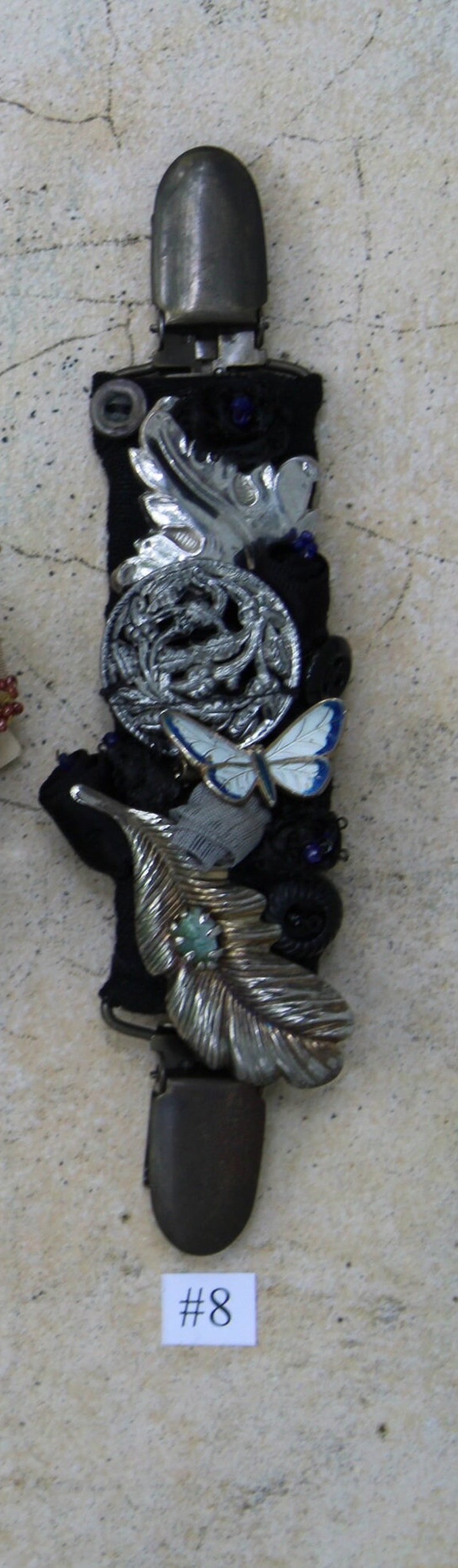 May include: A silver and black metal brooch with a butterfly, leaf, and floral design. The brooch is attached to a black fabric base. The number #8 is printed on a white label below the brooch.