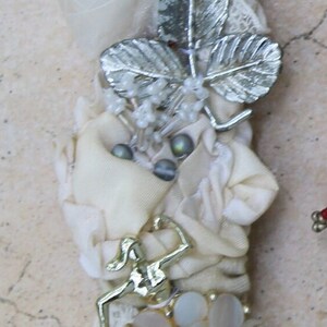 May include: A white fabric and lace embellishment with silver leaves, white beads, and a gold-toned figure. The embellishment is attached to a metal clasp with a silver finish. The embellishment is labeled #5.