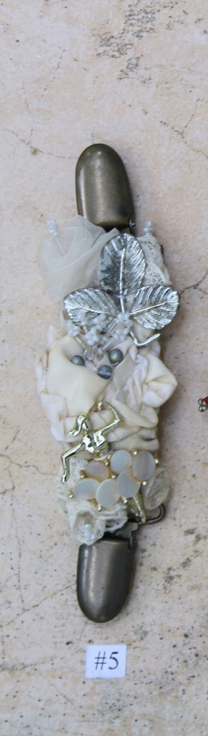 May include: A white fabric and lace embellishment with silver leaves, white beads, and a gold-toned figure. The embellishment is attached to a metal clasp with a silver finish. The embellishment is labeled #5.