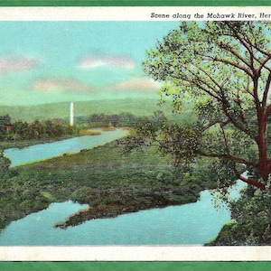 May include: A vintage postcard depicting a scenic view of the Mohawk River in Herkimer, New York. The postcard features a winding river with lush green foliage and a large tree in the foreground. The text "Scene along the Mohawk River, Herkimer, N.Y." is printed at the top of the postcard.