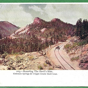 Puede incluir: Una postal antigua que representa una vía de tren serpenteando a través de un paisaje montañoso. El texto en la postal dice "103-Rounding The Devil's Slide. Colorado Springs & Cripple Creek Short Line."