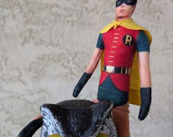 May include: A fabric doll of the character Robin wearing a red, yellow, and green costume with a black mask and yellow cape, positioned on top of a plastic owl figurine with large yellow eyes, black beak, and gray and white textured feathers.