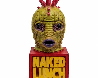 May include: A sculpted creature head with textured yellow-green skin, large dark eyes, and a purple mouth, topped with red protrusions. It sits on a red rectangular base featuring the yellow raised text NAKED LUNCH on the front face.