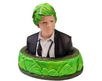 May include: A figurine bust of a person with bright green cabbage leaves for hair, wearing a black suit jacket over a white collared shirt with a dark tie. The base of the bust is circular and decorated with a sculpted ring of vibrant green cabbage leaves.
