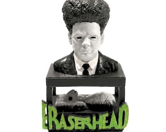 May include: A black and white bust of a figure with tall, textured hair, wearing a suit and tie. The bust sits on a black platform featuring a small, textured creature underneath and the word ERASERHEAD in bright green, raised lettering.