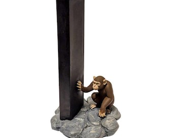 May include: A brown chimpanzee figurine crouched on a cluster of gray rocks, reaching out to touch a tall, vertical, rectangular black monolith. The sculpture is rendered in a realistic style with detailed fur texture and stone-like base.