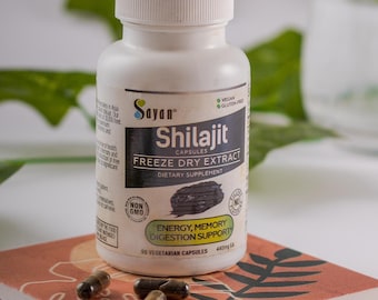 Puede incluir: Una botella blanca de cápsulas de suplemento dietético Sayan Shilajit Freeze Dry Extract. La botella está sobre una superficie con estampado rosa y marrón con varias cápsulas marrones esparcidas a su alrededor. La etiqueta de la botella dice "No GMO" y "Cápsulas vegetarianas".