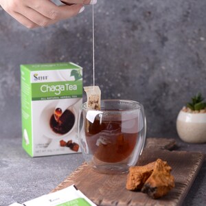 May include: A box of Chaga tea with a green and white label. The box is next to a glass cup of tea with a tea bag hanging from the rim. There are two tea bags on a wooden cutting board with a piece of chaga mushroom.