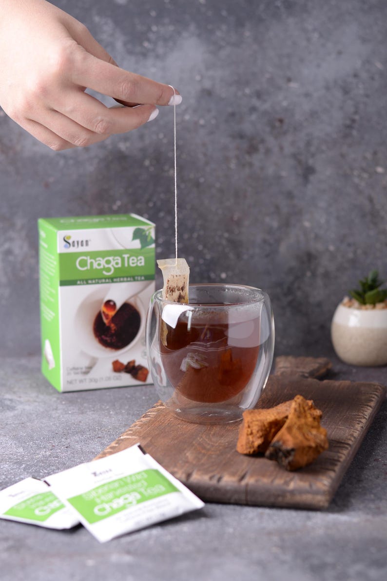 May include: A box of Chaga tea with a green and white label. The box is next to a glass cup of tea with a tea bag hanging from the rim. There are two tea bags on a wooden cutting board with a piece of chaga mushroom.