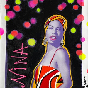 May include: A pop art style painting of a woman with the name "NINA" in pink script. The woman is wearing a colorful dress and headscarf, with a black background and colorful polka dots. The painting is framed in white.