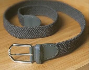 May include: A gray braided belt with a silver buckle. The belt is made of a woven material and has a leather finish.