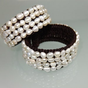 Puede incluir: Dos pulseras de crochet marrón con cuentas de perlas blancas. Las pulseras están apiladas una encima de la otra.