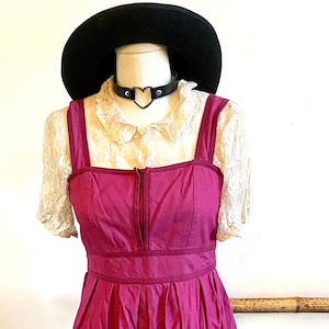 May include: A pink dress with a black choker and a black hat. The dress has a heart-shaped detail on the front and an apple design on the bottom. The dress is on a mannequin and the mannequin is standing on a blue wooden floor. There are two black boots with pink soles and silver chains on the floor.