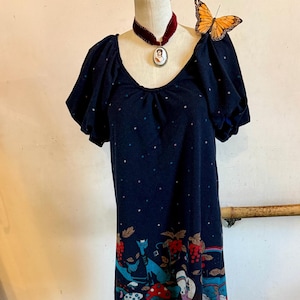 May include: Navy blue dress with a whimsical forest scene print at the hem. The dress features short sleeves and a scoop neckline. A velvet choker with a cameo pendant and a butterfly adorn the mannequin. White lace-up boots with black soles are at the base.