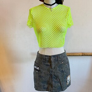 May include: A mannequin displays a neon green mesh top, a camouflage mini skirt with patches, and black platform boots. A plaid newsboy cap and a black choker with a cross pendant complete the outfit. The skirt has the letters "NIV" on a patch.