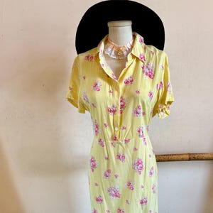 May include: A yellow floral print dress with pink flowers and short sleeves. The dress is displayed on a mannequin with a black hat and a decorative necklace. Black boots are at the base of the mannequin.