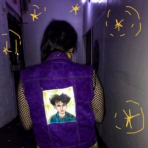 May include: A purple denim vest with a patch of a hand-painted portrait of a person with dark hair. The vest has a black fishnet shirt underneath and a plaid skirt.