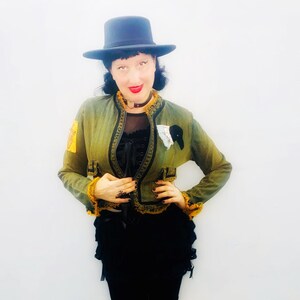 May include: A woman wearing a green jacket with gold trim, a black choker, and a black hat. She is standing in front of a white wall.
