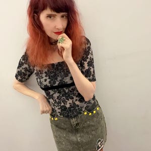 May include: A black floral lace top with short sleeves and a black velvet trim. The outfit includes a denim skirt with yellow stud accents and a patch. The person is wearing a black choker and a large green ring.