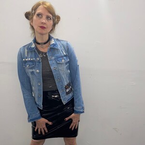 May include: A woman wearing a light blue denim jacket with distressed details, a black leather skirt, and black boots with silver studs. She is wearing a silver necklace and a black choker.