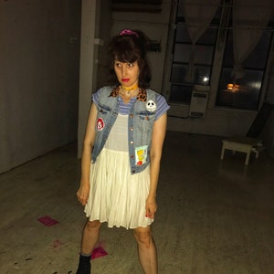 May include: A woman wearing a white dress with a denim vest over it. The vest has several patches, including a skull and crossbones, a cartoon character, and a red and yellow patch. She is also wearing a leopard print choker necklace and black boots.