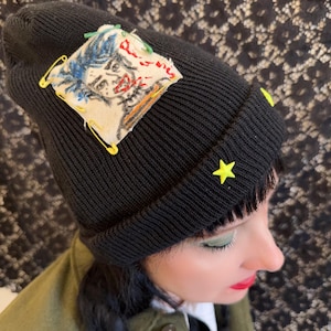 May include: Black knit beanie with a patch featuring a colorful, hand-drawn face design, secured with yellow safety pins. The beanie has two yellow star-shaped embellishments. The hat is worn by a person wearing an olive green jacket.