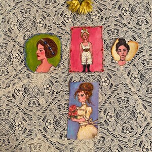 May include: Four small paintings on a white lace background. Each painting features a woman with a different hairstyle and outfit. The paintings are framed in a simple, white frame.