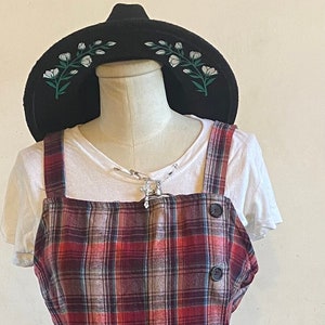 May include: A red, brown, and beige plaid overall dress with a white t-shirt underneath. The dress has a strawberry and bee patch on the front and black buttons down the side. The dress is on a mannequin with a black hat with white flowers on it. Black boots are on a platform below the mannequin.