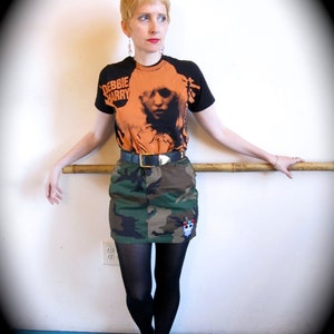 May include: A woman wearing a black and orange t-shirt with the text "Debbie Harry" printed on it. She is wearing a camouflage skirt, black tights, and black and white sneakers. She is standing in front of a white wall.