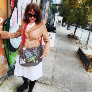 May include: A woman wearing a brown faux leather jacket with a fur collar, a white dress, and black knee-high boots. She is carrying a gray messenger bag with colorful patches that say "Skinny Puppy" and "Ministry".