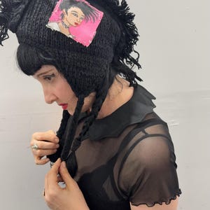 May include: Black knit hat with a mohawk-style fringe and braided ear flaps. A square patch on the hat features a portrait of a woman with pink background. The hat is a unique, handmade accessory.