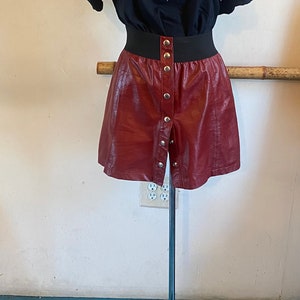 May include: A black t-shirt with a graphic print of a pumpkin and the text "HELLOWEEN are on 7 Sins". The shirt is paired with a burgundy faux leather skirt with button detailing and black platform shoes with red flames on the soles.