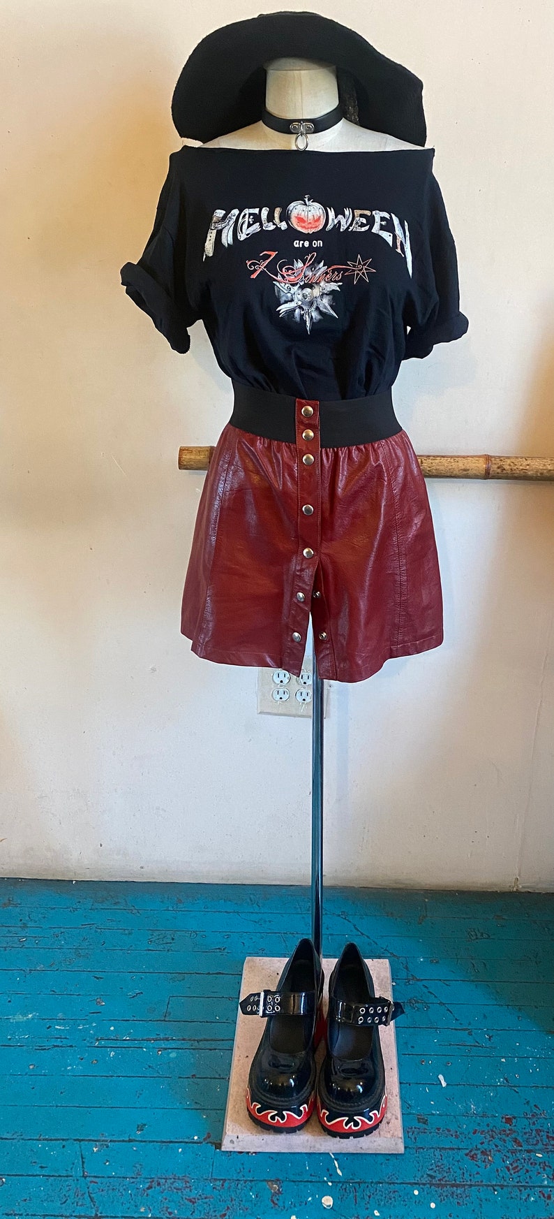 May include: A black t-shirt with a graphic print of a pumpkin and the text "HELLOWEEN are on 7 Sins". The shirt is paired with a burgundy faux leather skirt with button detailing and black platform shoes with red flames on the soles.