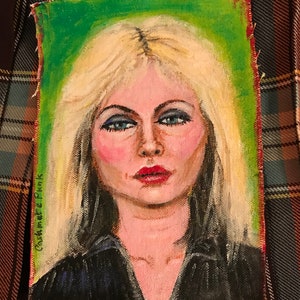 May include: A painting of a woman with blonde hair and blue eyes. She is wearing a black shirt and has a green background. The painting is signed "Catherine Pink".